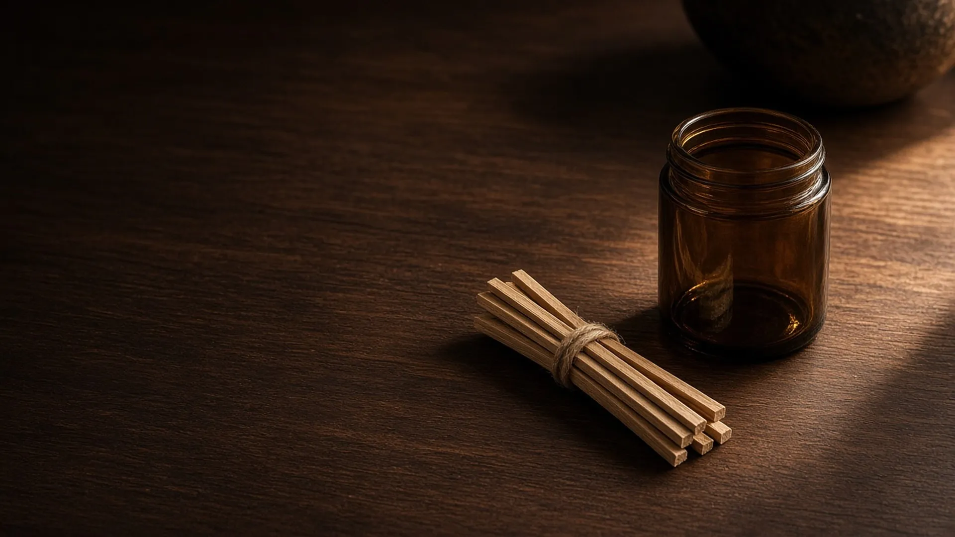 A bundle of thin wooden wicks tied with twine beside an empty amber jar on a walnut table