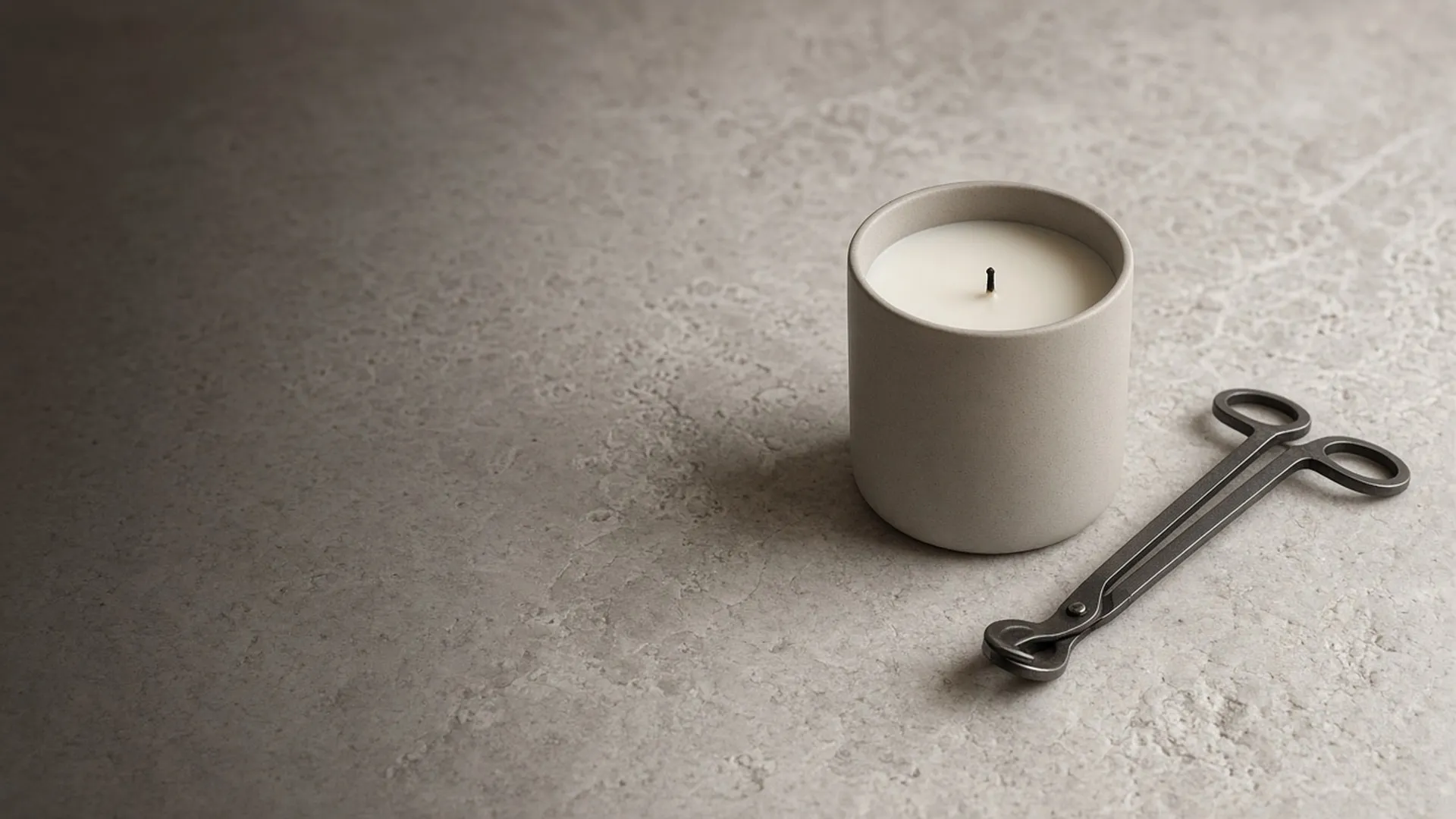 An unlit candle with a neatly trimmed wick and a pair of wick trimmers on a pale stone surface