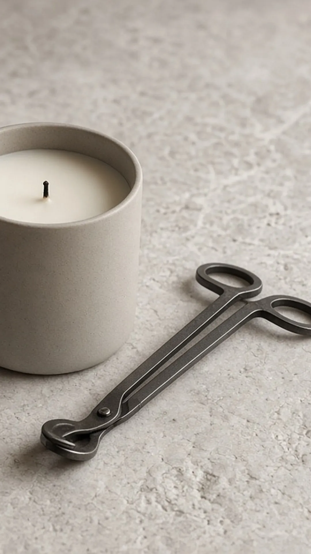 An unlit candle with a neatly trimmed wick and a pair of wick trimmers on a pale stone surface